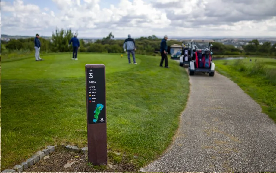 Vista geral de área de tee de campo de golfe com jogadores e foco no tee totem royal da floema em plástico reciclado castanho e HPL+.