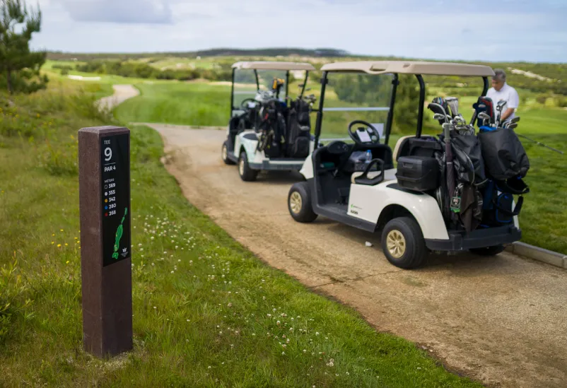 Tee totem Royal em campo de golfe para áreas de tee, produzido em plástico reciclado castanho e HPL+. Visíveis dois buggys de golfe com saco de tacos e um jogador.