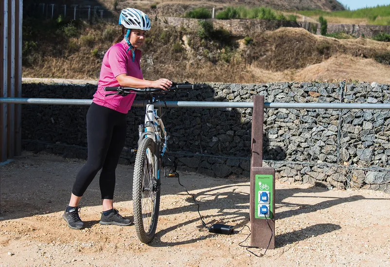 Person charging an electric bicycle using the Nature e-bike charger and Triatlo bicycle parking, Floema models, with recycled plastic frame, equipment for smooth electric mobility.