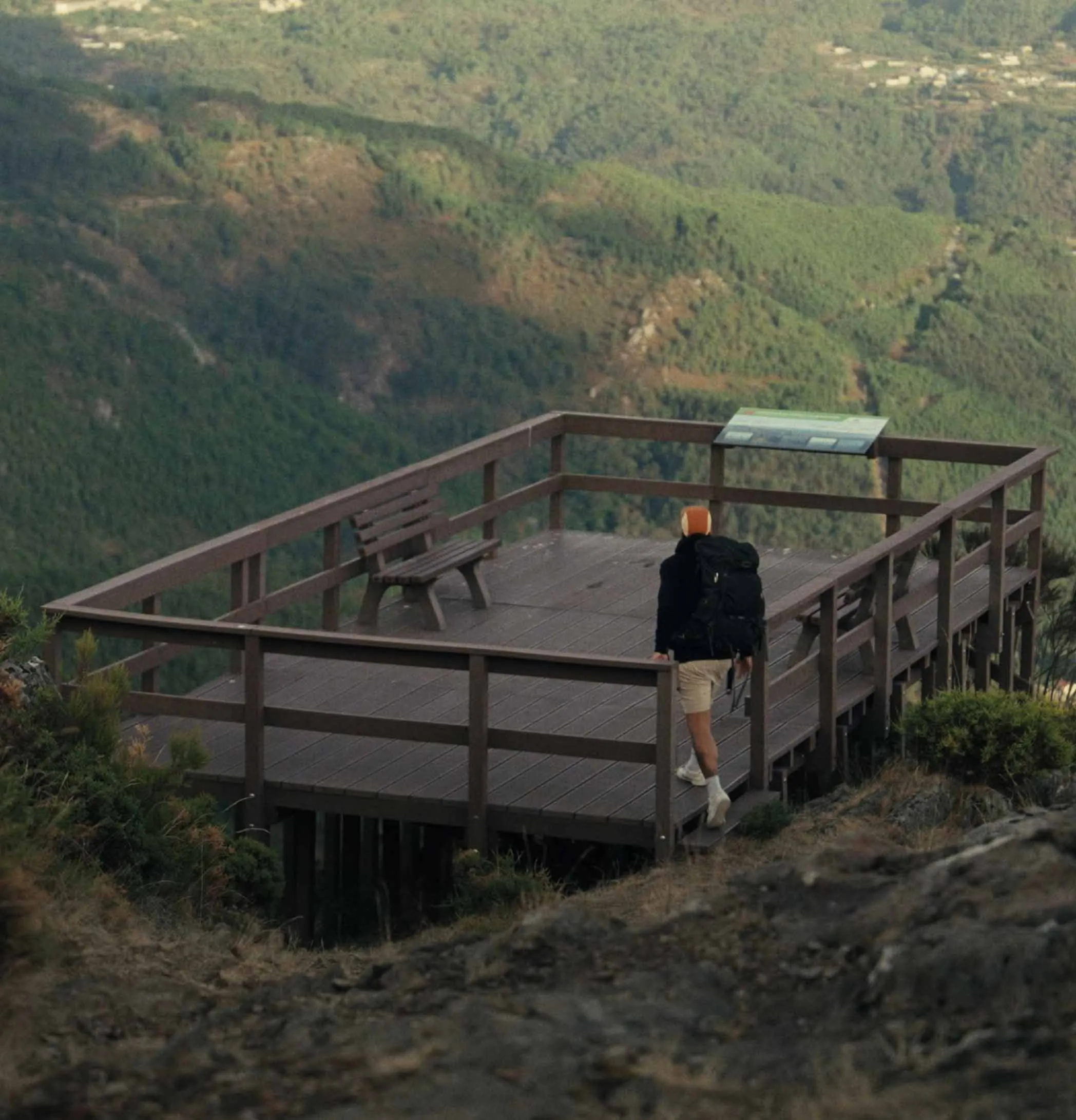 Caminhante a entrar em miradouro em plástico reciclado castanho Floema em espaço natural. Visível a montanha coberta de vegetação.